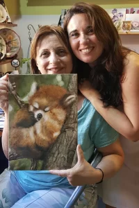 Marina Fiamingo smiling next to a student, both holding a hand-painted porcelain plaque with a realistic red panda, in her art-filled workshop.