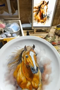 Porcelain plate with realistic portrait of a horse, paintbrushes and tablet with visual reference on a wooden table, illustrating the discipline of observation in Marina Fiamingo's workshop.
