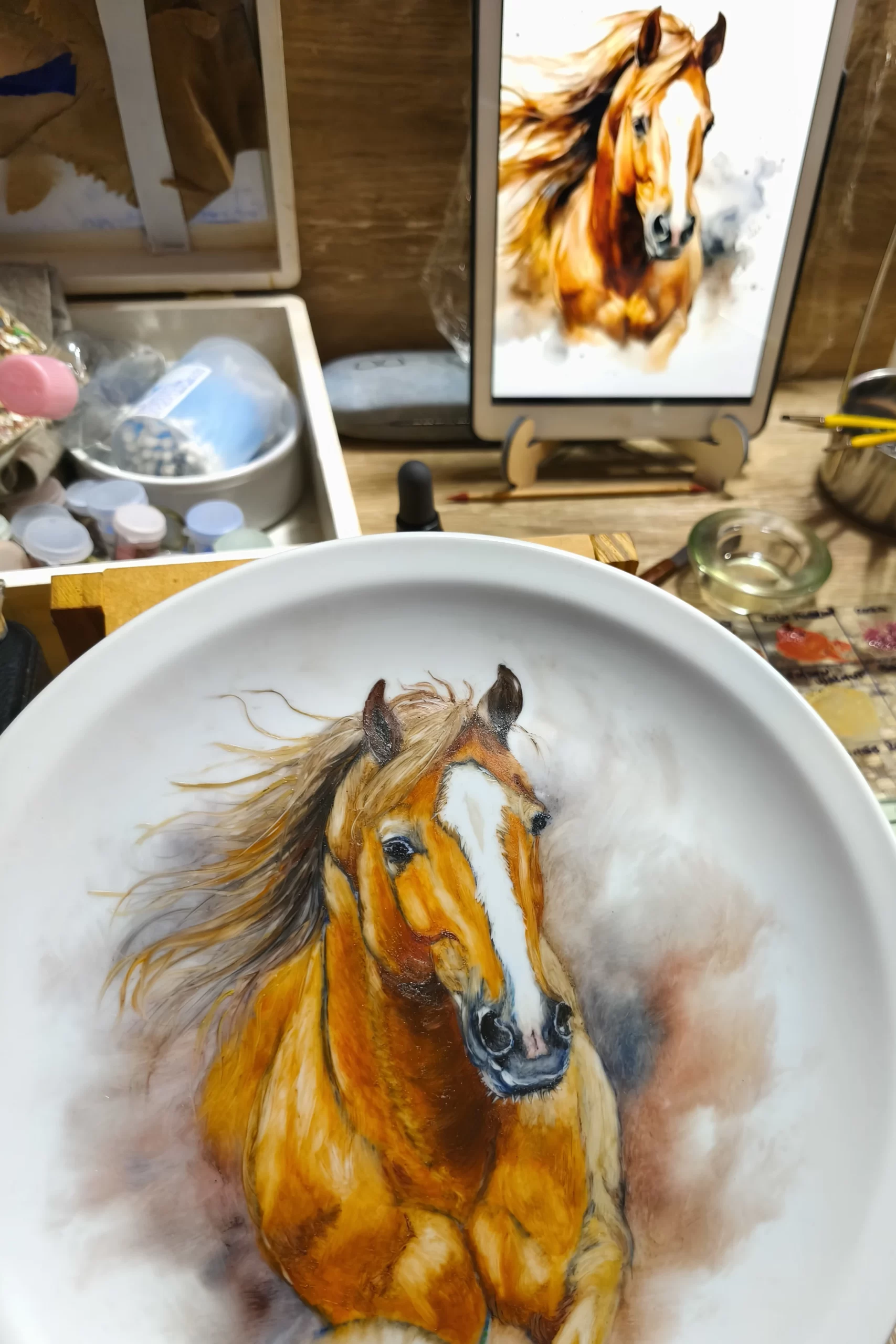 Porcelain plate with realistic portrait of a horse, paintbrushes and tablet with visual reference on a wooden table, illustrating the discipline of observation in Marina Fiamingo's workshop.